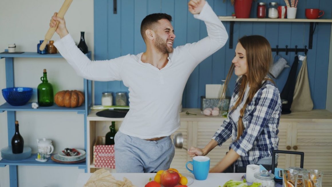 Attractive young joyful couple have fun dancing and singing while set the table for breakfast in the kitchen at home