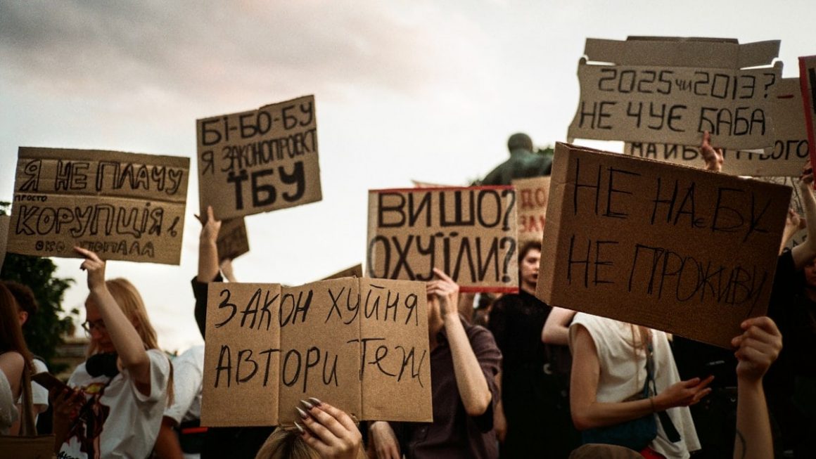 Protesters hold signs with handwritten messages against a cloudy sky.